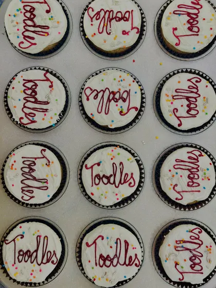 A tray of our vanilla buttercream cupcakes decorated for a corporate farewell party. Each cupcake is topped with the fun message "Toodles" and colorful sprinkles for a sweet and cheerful send-off.