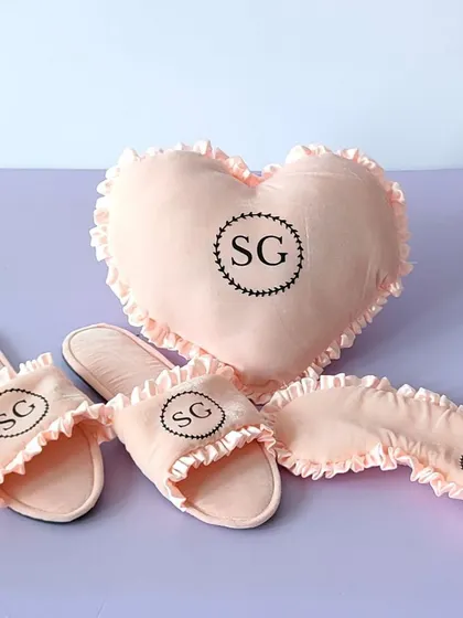 A cozy and personalized gift set for a bridesmaid, including a heart-shaped pillow, slippers, and an eye mask, all customized with her initials.