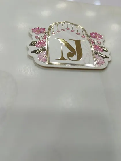 A close-up of a personalized gold tag with the couple's initials, set against a beautiful Pichwai-themed design with lotus and jharokha details.