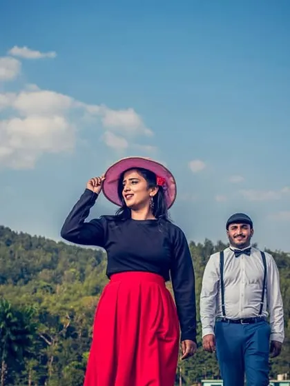 A stylishly dressed couple walks through a green landscape under a bright blue sky. This image has a classic, almost vintage feel, perfect for a timeless pre-wedding album.