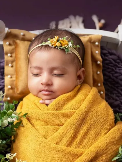 A 27-day-old baby girl wrapped in yellow, sleeping on a purple blanket with floral accents.