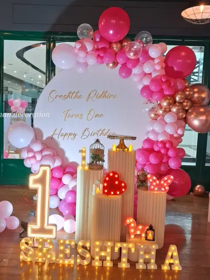 A lovely pink and rose gold theme for a first birthday. The setup includes a circular backdrop, an elaborate balloon arch, marquee letters, and multiple pedestals with light-up props.