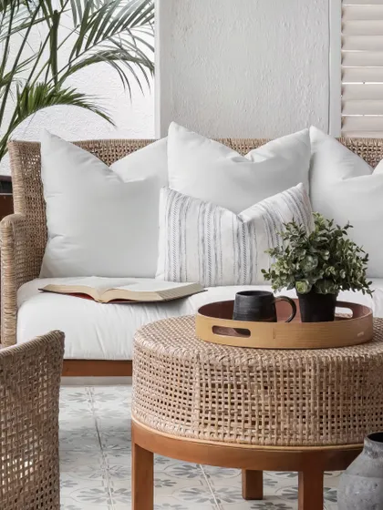 The rattan coffee table and sofa, adorned with soft white cushions and a simple wooden tray, create a serene and comfortable setting. The patterned floor tiles add a subtle decorative touch.