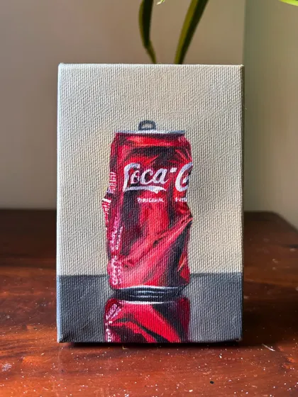 The finished oil painting of the crushed Coke can, sitting on a wooden table. The reflection on the surface below adds another layer of realism to the piece.