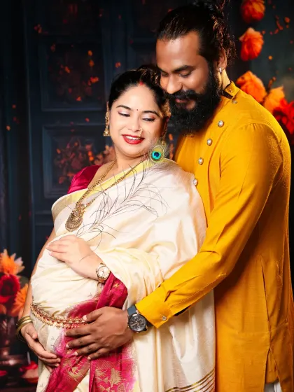 A close-up shot filled with emotion. The couple shares a tender moment, their hands resting on the baby bump, set against a backdrop rich with traditional Indian decor.