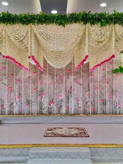 This stage was designed for a modern Janoi ceremony with a serene pink and white color palette. The backdrop is a curtain of delicate white floral strings accented with pink flowers, framed by traditional banana trees, blending tradition with contemporary aesthetics.