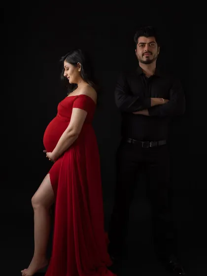 A stylish and confident pose from a couple's maternity session. The mother-to-be stands in profile showcasing her bump in a red gown, while her partner stands behind her with a protective stance against a black background.