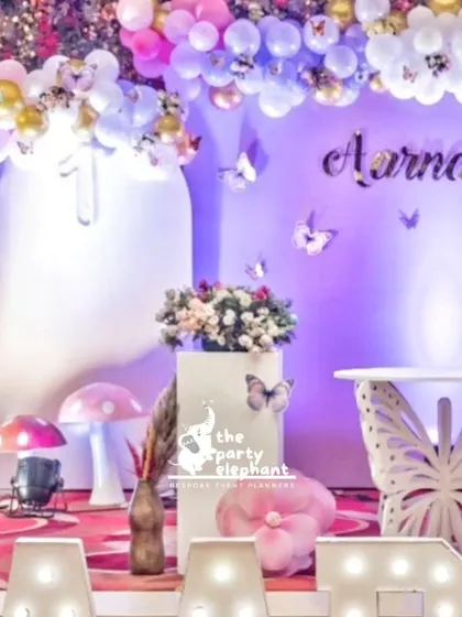 The main stage is beautifully lit, highlighting the personalized name, butterfly table, and surrounding floral and balloon arrangements.