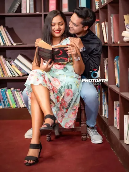 A romantic library-themed pre-wedding photo. The groom embraces his partner from behind as she reads a book, creating a sweet, shared moment.