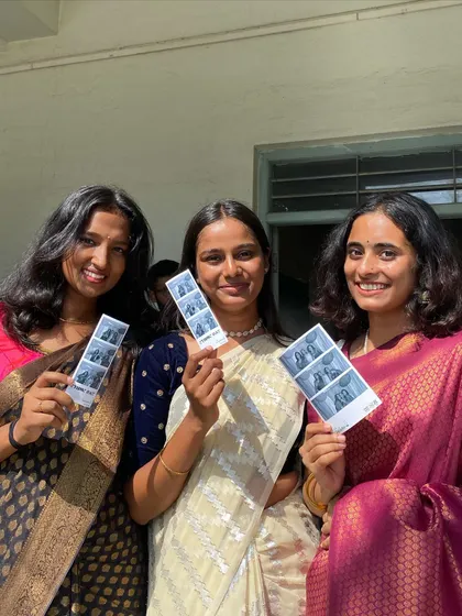 Three friends in elegant sarees, each holding a photo strip from the event.