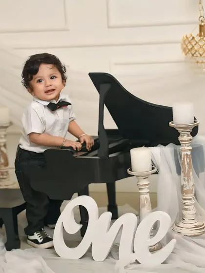 A handsome little gentleman dressed in a bowtie stands next to a miniature piano for his first birthday photoshoot, a creative and elegant way to mark the milestone.