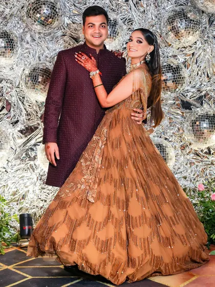 A full-length portrait of the couple at their sangeet. The bride's flowing gown and the groom's elegant attire are perfectly complemented by the shimmering, festive background.