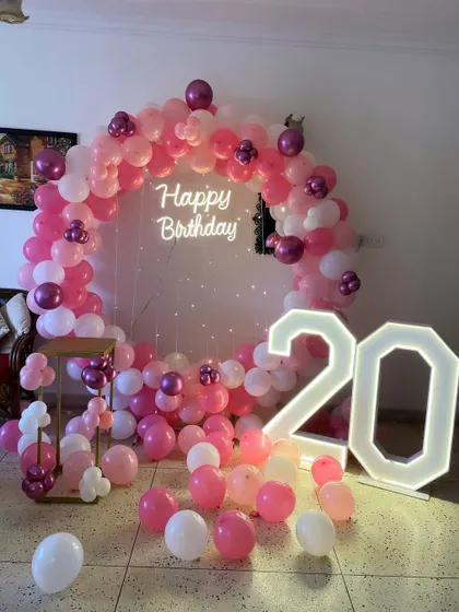 A pretty pink-themed 20th birthday. This setup includes a circular balloon arch, a neon sign, and large light-up numbers, with balloons scattered on the floor.