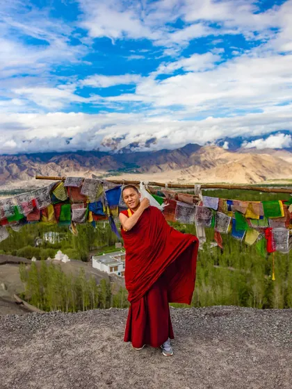 A Buddhist nun in red robes stands on a hilltop in Ladakh, with prayer flags and the vast mountain range behind her. The image conveys a sense of freedom and spiritual devotion.