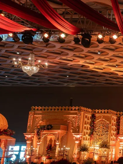 A partial view of a grand, temple-inspired wedding stage with red fabric draping and crystal chandeliers, showcasing the scale and architectural detail of the design.