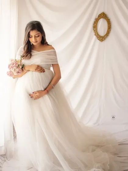 A full-length shot of a mother-to-be in a flowing white gown, capturing a moment of quiet contemplation by the window.