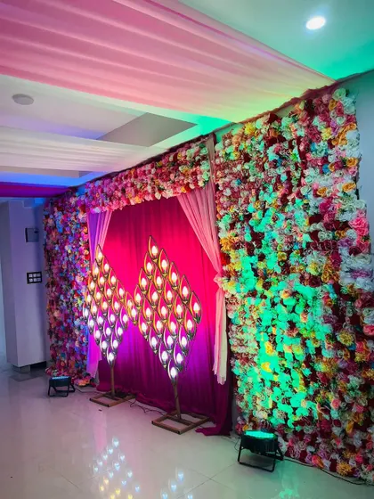A vibrant stage backdrop for a Sangeet or reception. This setup features a wall of colorful flowers and unique peacock-feather-shaped lamps.