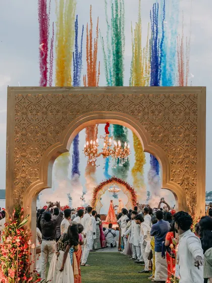 A wide shot of the wedding aisle, showing the grand scale of the celebration as guests witness the varmala against a backdrop of colorful smoke.