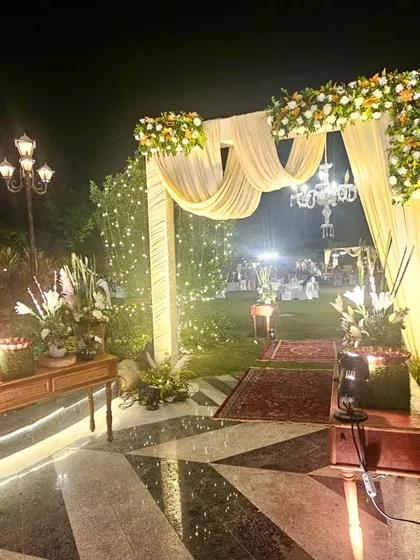 An elegant outdoor engagement ceremony entrance at night. The archway is draped in soft cream fabric and adorned with white and orange flowers, with a chandelier adding a touch of sparkle.
