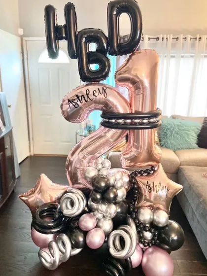 A personalized balloon bouquet for a 21st birthday. This large arrangement features number balloons, a custom name, and a color scheme of rose gold, black, and silver.