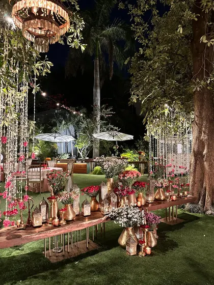 A long table decorated with an abundance of flowers and brass lanterns, under hanging chandeliers.