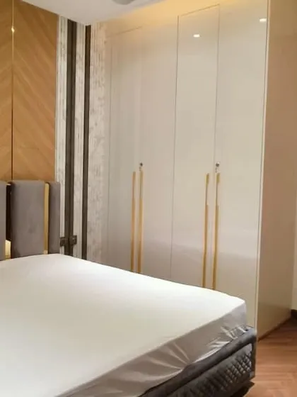A modern luxury bedroom featuring a high-gloss white wardrobe with long gold handles. The accent wall behind the bed combines geometric wood paneling with marble for a plush look.