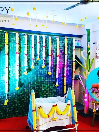 A charming cradle ceremony setup for a home event. The backdrop is made of woven coconut leaves, decorated with flower garlands, and personalized with a photo of the baby.