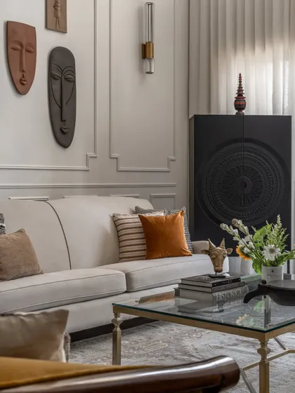 A close-up of the living room decor, featuring a glass coffee table styled with books and flowers. The carved black cabinet and wall of masks in the background add to the eclectic, collected feel of the home.