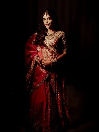 A full-length portrait against a dark background, emphasizing the grandeur of the red bridal lehenga. The lighting focuses solely on the bride, making her the center of this artistic shot.