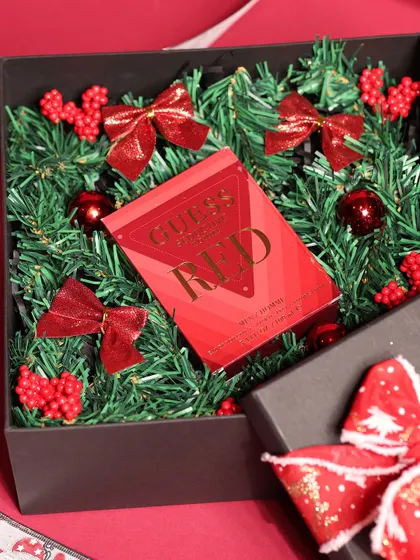 A Christmas gift for him featuring a Guess Seductive Red perfume, presented in a box filled with festive greenery and red bows.