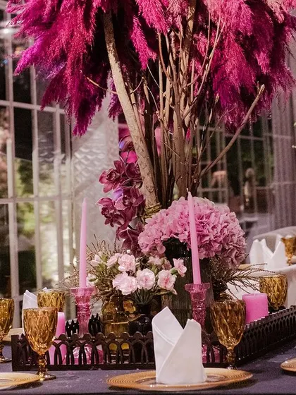 A stunning tablescape from a chic pre-wedding event. The centerpiece features dramatic pink pampas grass and hydrangeas, set against amber-colored glassware and gold plates.