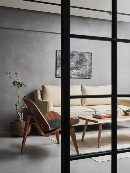 A view into the living room through the glass partition, showcasing the simple, curated furniture and the textural concrete walls. This approach creates layers and depth within a minimalist framework.