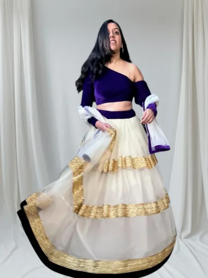 A modern and chic lehenga choice. This outfit features a rich purple velvet drop-shoulder blouse paired with a tiered ivory and gold skirt, perfect for a cocktail party or reception.