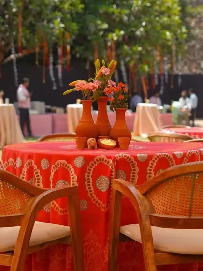 A close up of a guest table setting. The combination of the patterned red cloth, wooden chairs, and a simple terracotta vase centerpiece creates a look that is both rustic and culturally rich.
