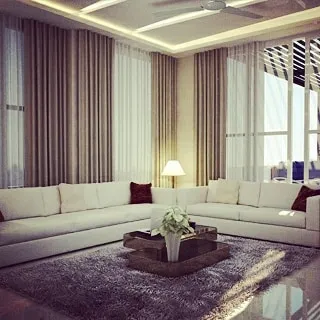 This living room design emphasizes comfort and light. The large sectional sofa, plush area rug, and floor-to-ceiling curtains create a soft and inviting space for the family to gather.