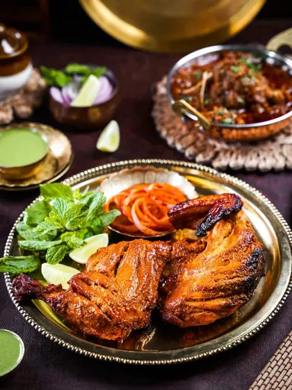 A classic Tandoori Chicken served on a brass plate with mint chutney and onion salad. The rustic, dark styling highlights the charred texture and vibrant red color of the dish.