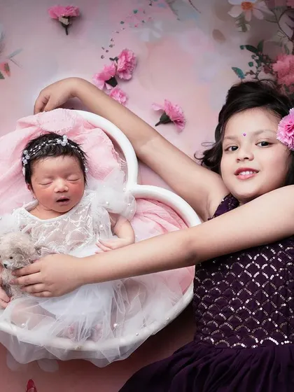 A beautifully styled sibling portrait. The older sister proudly presents her sleeping baby sister in a heart-shaped basket, surrounded by flowers.