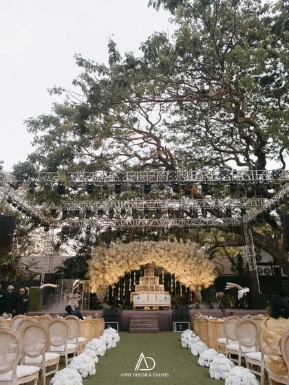 A wide shot of a grand outdoor sangeet, showing the guest seating arrangement with a green aisle leading to a magnificent stage framed by a large tree and professional lighting rigs.