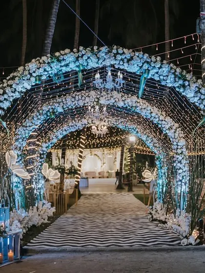 A magical "under the sea" or "winter wonderland" themed entrance tunnel. The arch is covered in white flowers and illuminated with blue lights and fairy light curtains for a truly immersive experience.
