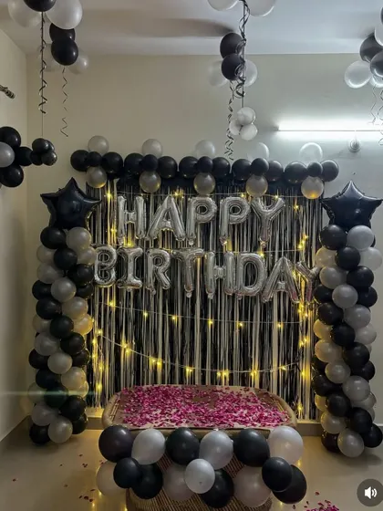 A birthday setup with a black shimmer curtain, silver foil letters, and balloon pillars. The table is covered with rose petals for a special touch.