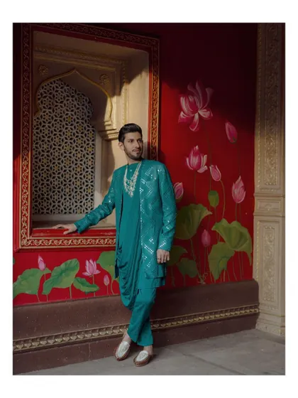 A stylish portrait of the groom in a unique teal kurta set, posing against a vibrant red wall with lotus murals.