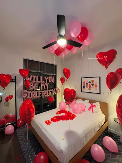 I can help you ask the big question with a beautiful surprise setup. This room features 'WILL YOU BE MY GIRLFRIEND' foil balloons, a mix of red and pink latex and heart-shaped foil balloons, and a bed covered in rose petals to create the perfect romantic proposal setting.
