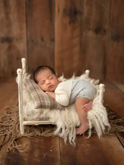 A full-length view of a newborn boy sleeping on a miniature bed. The rustic wooden floor and fluffy rug complete this charming setup.