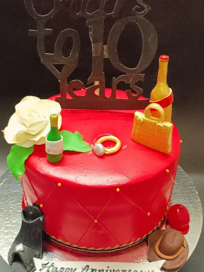 A vibrant red cake for a 10-year anniversary, decorated with edible wine bottles, a ring, and a "Cheers to 10 Years" topper.