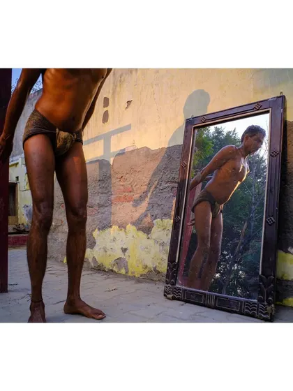 A wrestler looks at his reflection in a mirror propped against a wall at an outdoor gym. The golden hour light and the subject's pose create a moment of introspection and pride.
