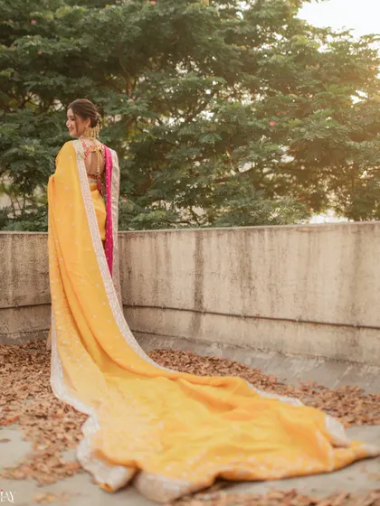 This shot from behind showcases the dramatic and beautiful trail of the bride's yellow nauvari saree against a bed of autumn leaves. It highlights the elegance and grandeur of the traditional Maharashtrian bridal look.