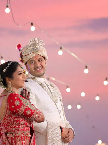 A romantic shot of the couple against a beautiful pink and purple sunset sky. The string lights add a magical touch to this candid wedding portrait.