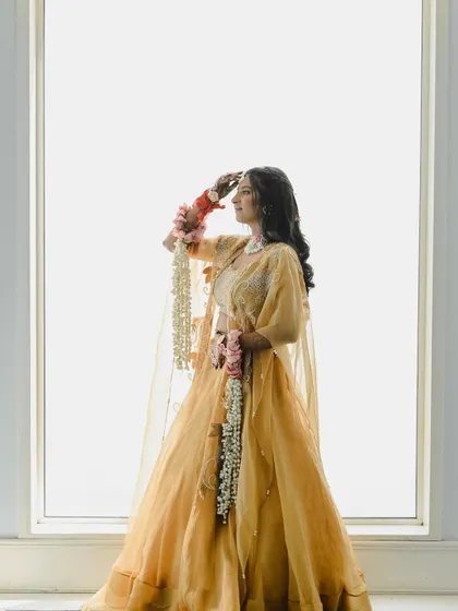 A high-key portrait against a white background, creating a soft and ethereal look. The bride, holding her floral kaleeras, looks like a vision in her golden-yellow lehenga.