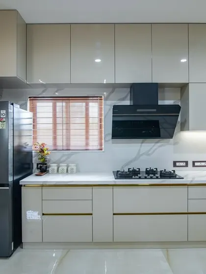A transformed kitchen with sleek, light grey glossy cabinets and gold-accented handles. The design includes a marble-look backsplash, a black chimney, and a side-by-side refrigerator for a modern, high-end finish.