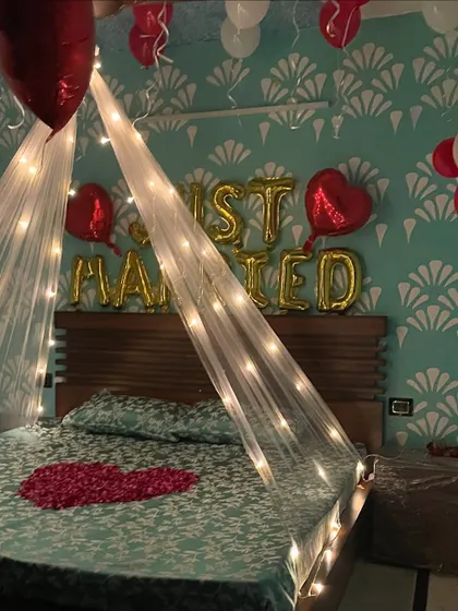 A romantic 'Just Married' room setup. The decor features a bed canopy with fairy lights, red and white balloons, and a heart made of rose petals on the bed.